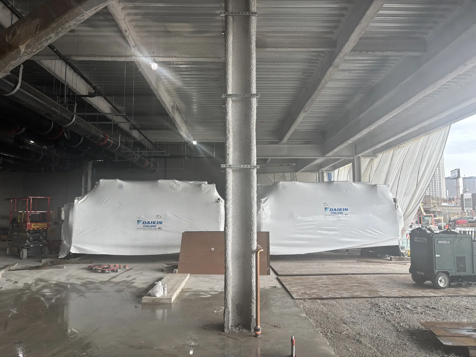 Two large shrink-wrapped air handling units staged under a concrete deck on a construction floor