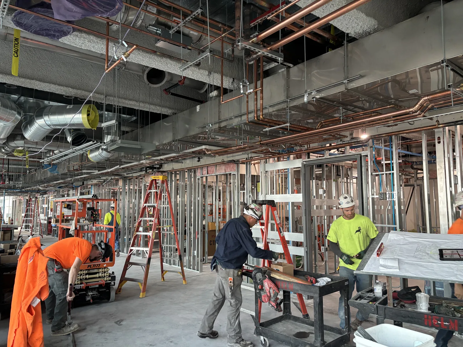 A commercial interior under construction with ladders, sawhorses, exposed overhead services and several trades at work