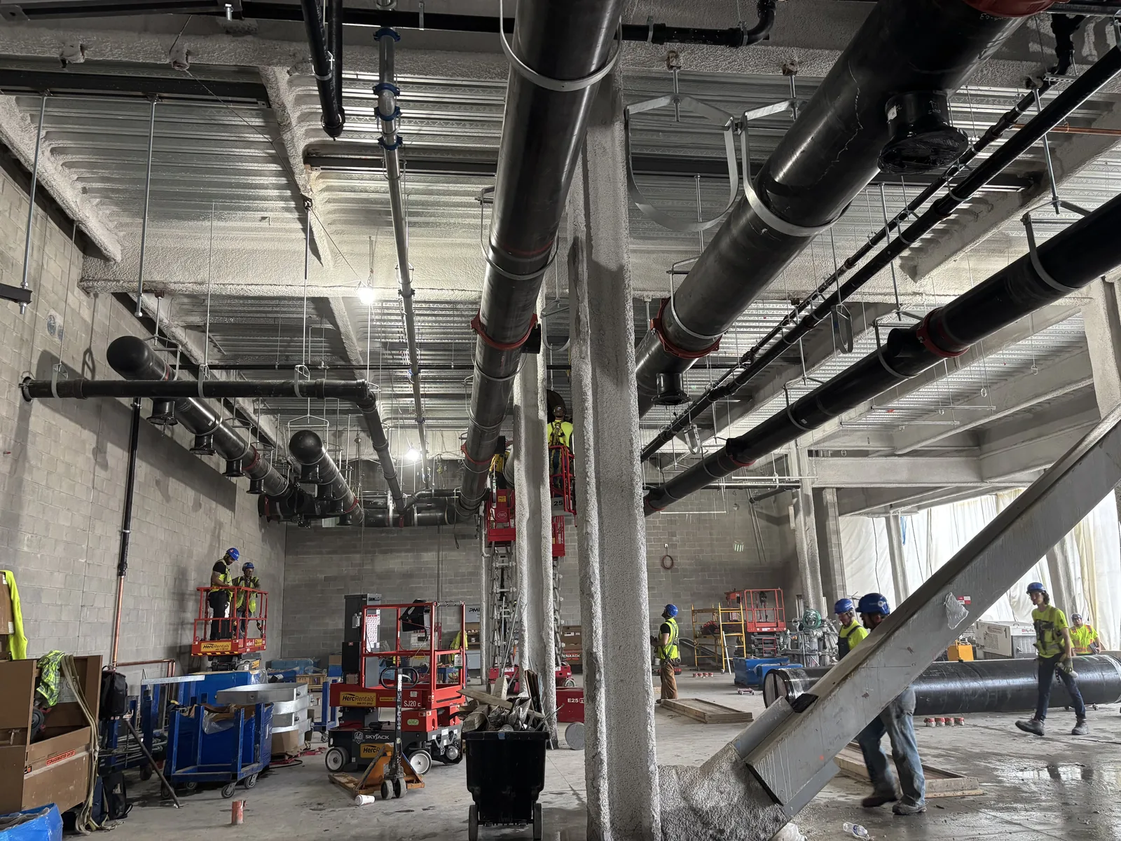 Overhead ductwork and pipe runs in a commercial building mid-fit-out with trades working in the background