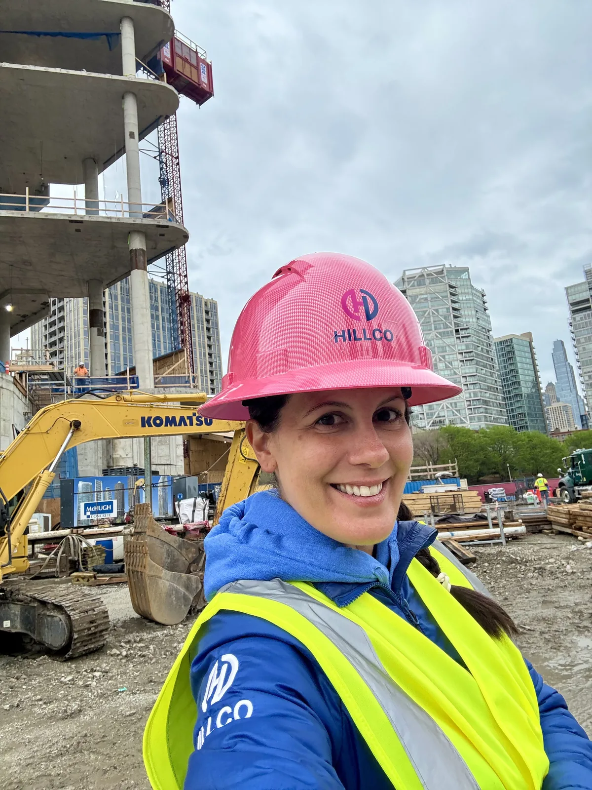 Nikki Liddy in a pink Hillco hard hat and safety vest on a jobsite with an excavator and high-rise buildings behind her