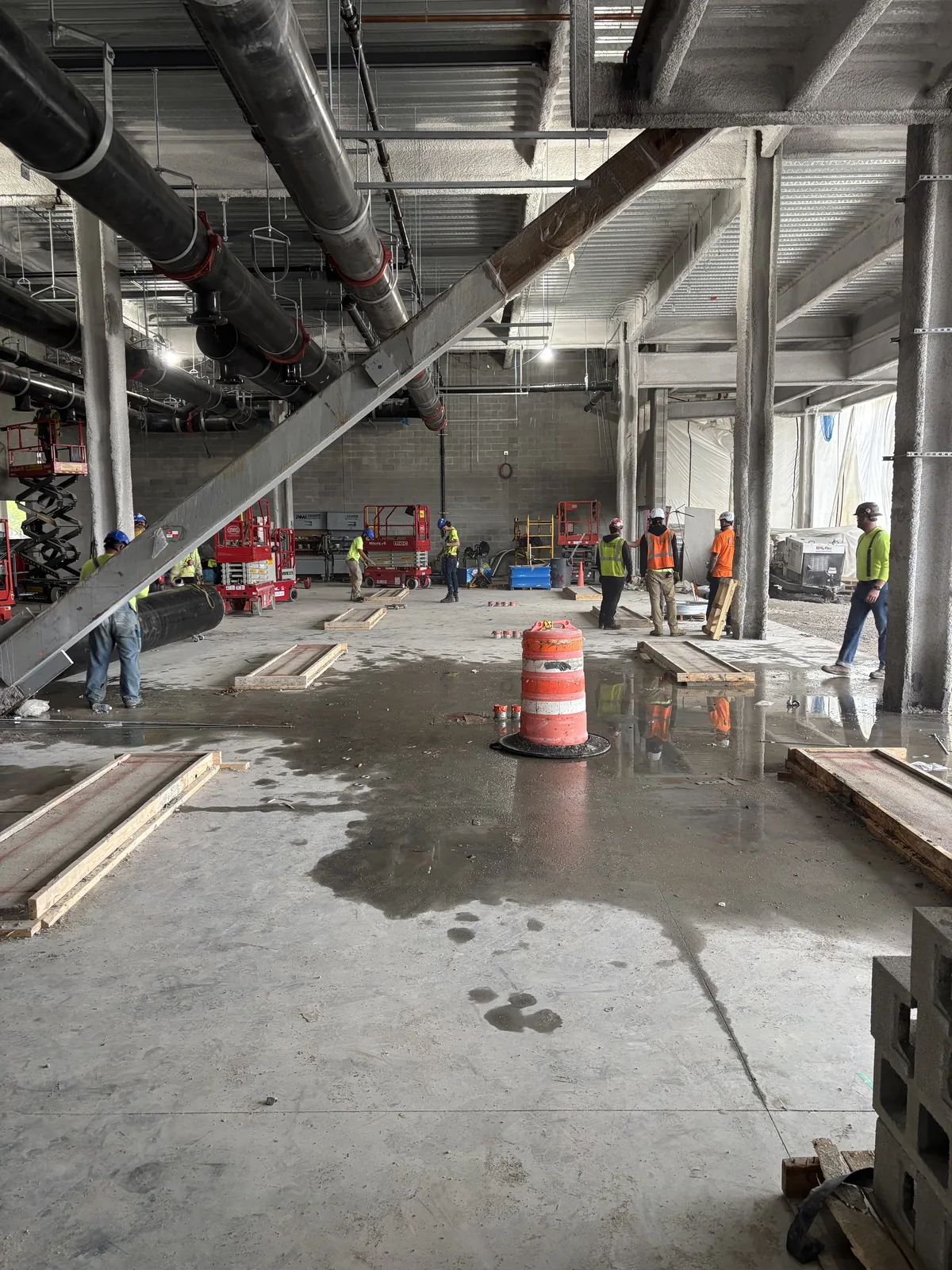An open concrete floor mid-build with an orange riser sleeve, overhead piping, and tradespeople working