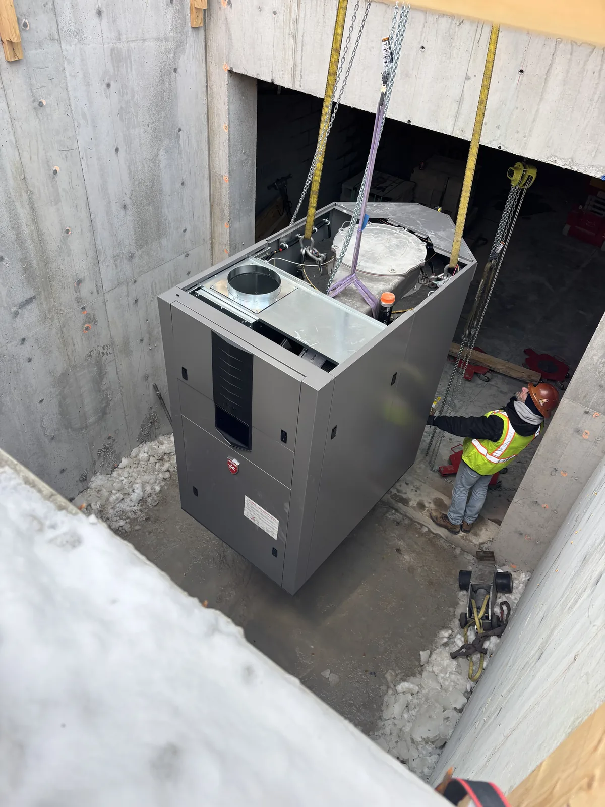 A packaged HVAC unit in rigging straps being lowered through an opening cut in a concrete slab, with a worker in a high-visibility vest guiding it